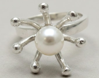 May include: A silver ring featuring a central white pearl surrounded by six radiating silver prongs, each topped with a small polished silver sphere. The band is smooth and metallic, creating a starburst effect around the round white pearl.