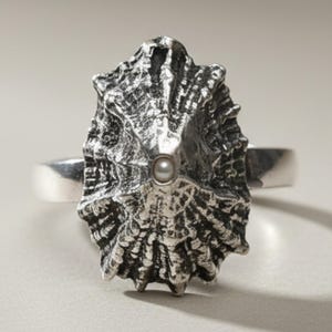 May include: A silver ring featuring a textured, conical shell-shaped centerpiece with a small, round white pearl embedded at the apex. The metal surface has a dark, oxidized finish that highlights the intricate, radial ridges of the shell design.