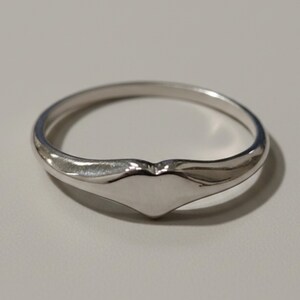 May include: A polished silver-toned ring featuring a smooth, solid band that tapers into a subtle, raised heart-shaped focal point at the center. The metal surface is reflective and uniform, showcasing a clean, minimalist design with no additional stones.
