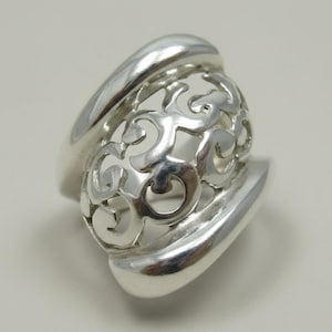 Puede incluir: Un anillo de plata pulida con una banda ancha y lisa que se transforma en una secci&oacute;n central con un intrincado patr&oacute;n de filigrana calada. El dise&ntilde;o consiste en bucles met&aacute;licos entrelazados y giratorios que crean una estructura hueca.