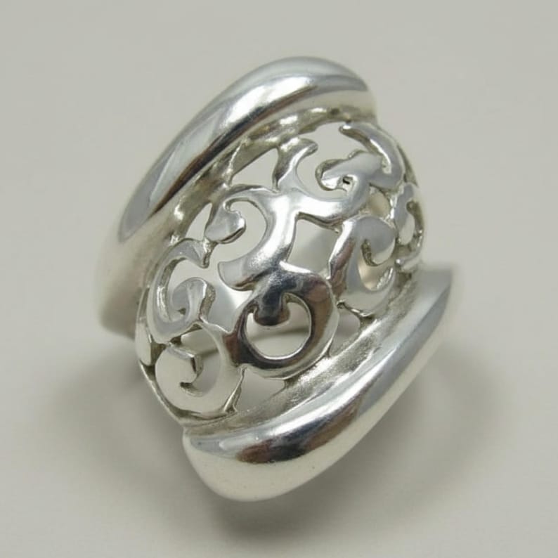 Puede incluir: Un anillo de plata pulida con una banda ancha y lisa que se transforma en una secci&oacute;n central con un intrincado patr&oacute;n de filigrana calada. El dise&ntilde;o consiste en bucles met&aacute;licos entrelazados y giratorios que crean una estructura hueca.