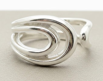 May include: A polished silver ring featuring an open, bypass-style design with two concentric, rounded oval loops that overlap. The band is smooth and reflective, showcasing a continuous, fluid metallic form that wraps around the finger.