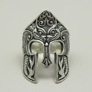 Op de afbeelding: Een zilverkleurige metalen ring in de vorm van een spartaans helm. Het oppervlak is voorzien van gedetailleerde gravures, waaronder gestileerde vlammenpatronen en symmetrische krulmotieven. Het ontwerp bevat een centrale neusbeschermer en twee zijplaten.