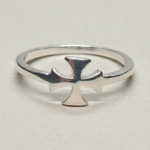 Könnte beinhalten: Ein polierter Silberring mit einem mittig platzierten Kreuzemblem. Die Ringschiene ist glatt und kreisförmig und geht nahtlos in die ausgestellten Arme des Kreuzdesigns über. Das Metall weist durchgehend eine reflektierende Oberfläche auf.