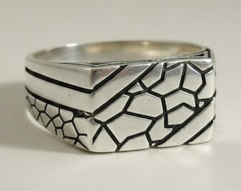 May include: A silver ring featuring a flat, rectangular top surface with a geometric, cracked-pattern design. The band is polished silver with deep, dark recessed lines that create a segmented, mosaic-like texture across the entire front face.