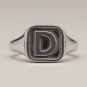 Op de afbeelding: Een zilverkleurige zegelring met een vierkant vlak met afgeronde hoeken. Het midden van de ring toont een prominente, verhoogde hoofdletter D in een vet, schreefloos lettertype, geplaatst tegen een verzonken, donkere, geoxideerde achtergrond.