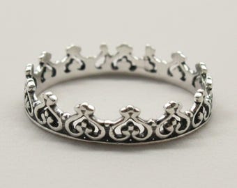 May include: A silver ring shaped like a crown, featuring an intricate openwork design with repeating heart-like motifs and pointed peaks along the top edge. The metal has a polished finish with darkened recessed areas to highlight the detailed pattern.