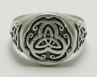 May include: A silver-toned metal ring featuring a circular face with an embossed triquetra knot design. The band displays matching intricate knotwork patterns on both sides, with darkened recessed areas that highlight the raised metal details.