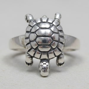 May include: A silver ring featuring a three-dimensional turtle design on the front. The turtle shell has a segmented, rounded pattern with oxidized detailing in the grooves, while the head and four legs extend from the central shell element.