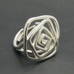 May include: Silver ring with a geometric design resembling a rose. The ring is made of a single piece of metal that is twisted and shaped into a complex pattern.
