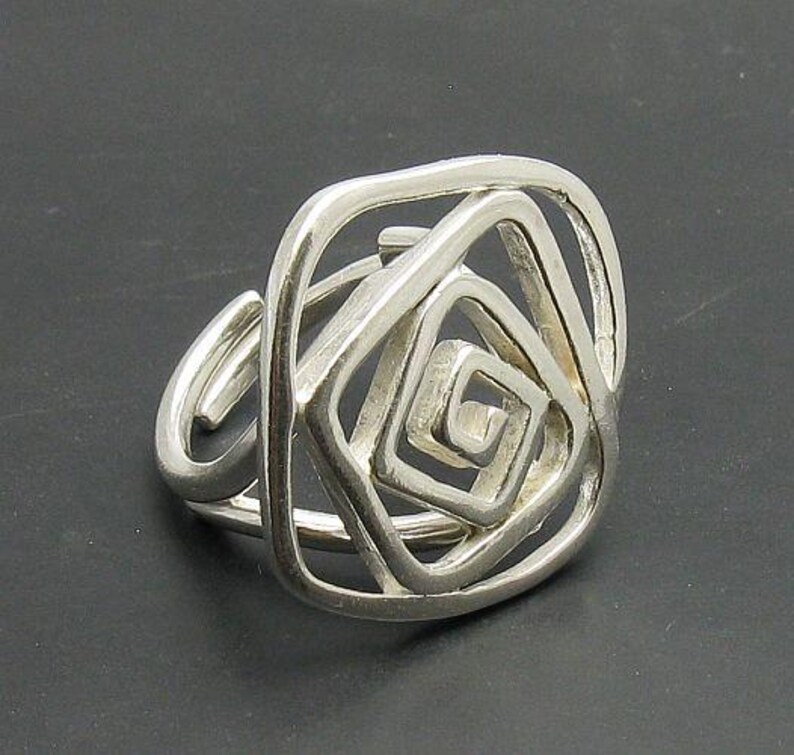 May include: Silver ring with a geometric design resembling a rose. The ring is made of a single piece of metal that is twisted and shaped into a complex pattern.