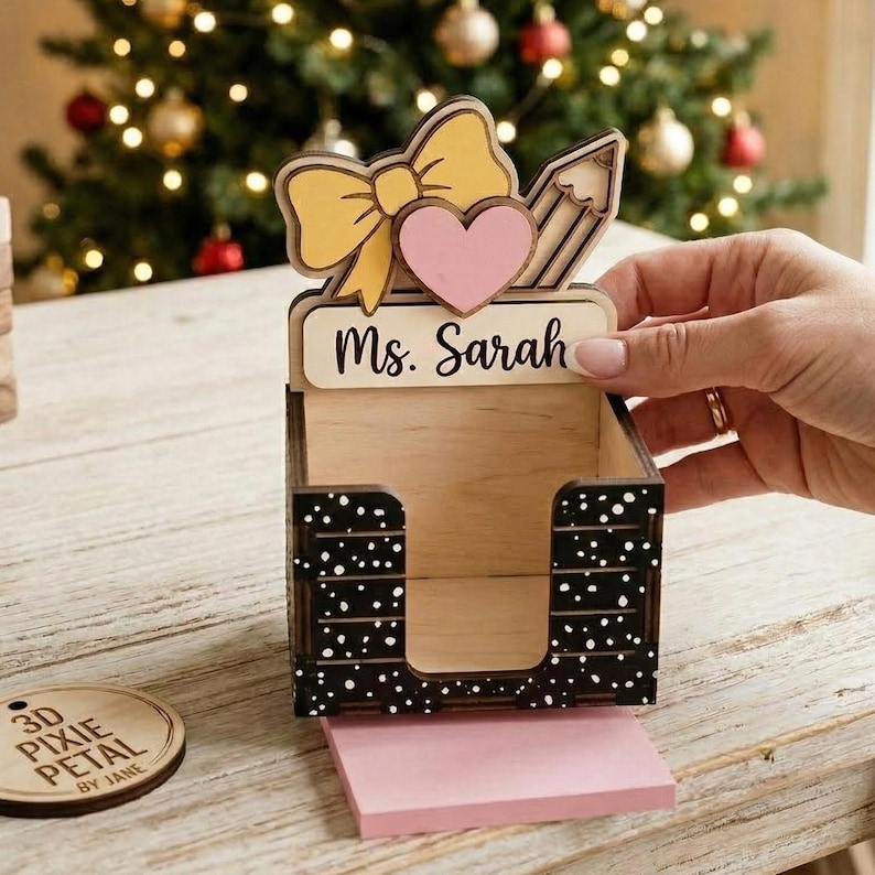 May include: A wooden desk organizer with a bow, heart, and pencil design. The organizer has the name "Ms. Sarah" on it and a black and white polka dot pattern. A stack of pink sticky notes is visible.