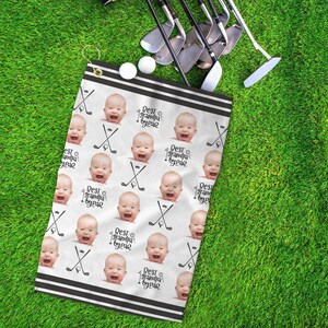 Op de afbeelding: Witte golfhanddoek met een zwarte rand en herhaalde afbeeldingen van een babygezicht met de tekst "Best Grandpa by Year" erop gedrukt.