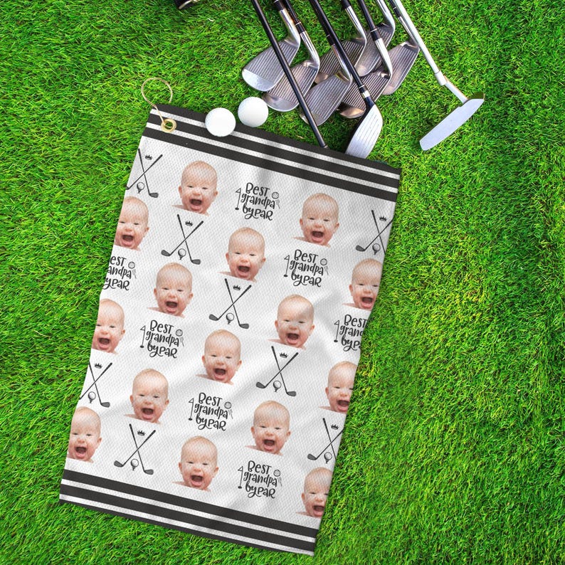 Op de afbeelding: Witte golfhanddoek met een zwarte rand en herhaalde afbeeldingen van een babygezicht met de tekst "Best Grandpa by Year" erop gedrukt.