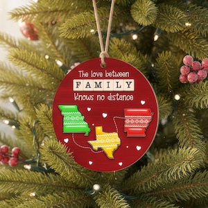 May include: A round, red Christmas ornament with the text "The love between FAMILY Knows no distance." The ornament features green, yellow, and red state outlines with white heart accents. A beige string is attached for hanging.