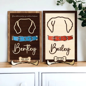 May include: Two wooden plaques with dog silhouettes, collars, and names. The plaques are brown with a light brown bone shape at the bottom. The text on the plaques reads "Bentley 2012-2024" and "Bailey 2012-2024".