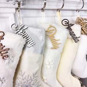 May include: Five white Christmas stockings with personalized wooden name tags. The stockings are decorated with fur, glitter, and pom-poms. The names on the tags are Charlie, Mommy, Daddy, Amanda, and Ariana.
