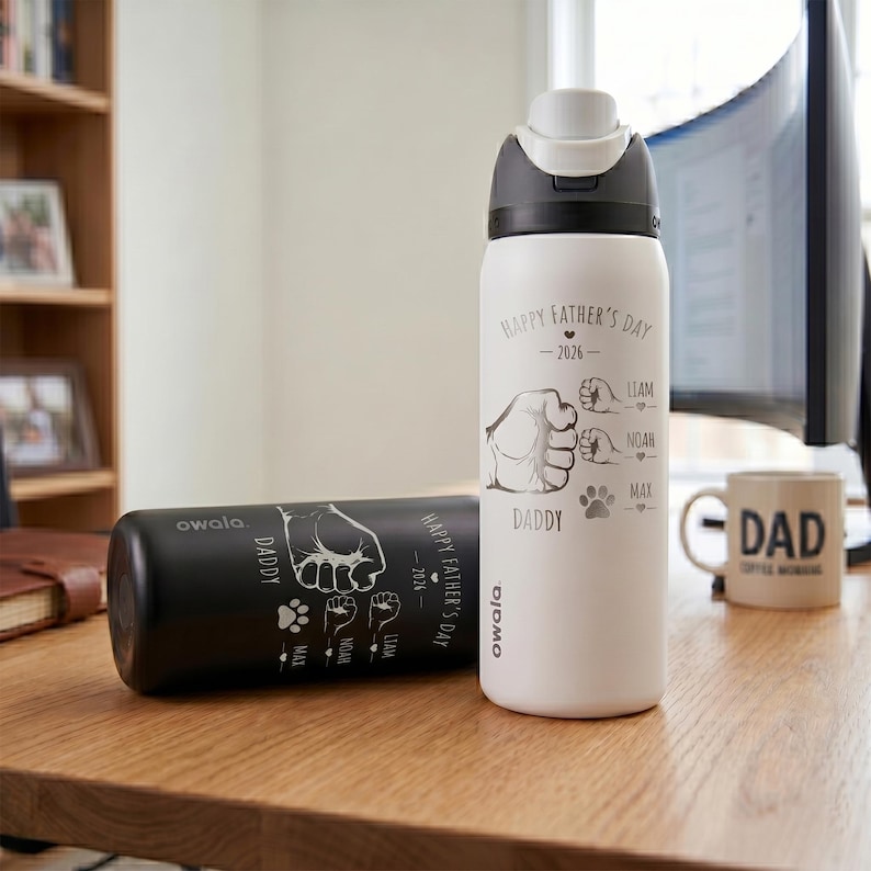 May include: Two insulated water bottles, one black and one white, with a flip-top lid. The white bottle has "Happy Father's Day - 2026" and "Daddy" printed on it, along with fist and paw print graphics. The black bottle has similar text and graphics.