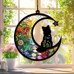 Puede incluir: Un carill&oacute;n de viento en forma de luna de vidrio tintado con una silueta de un perro sentado en la luna creciente. El vidrio presenta un dise&ntilde;o floral colorido con una salida del sol en el fondo. El nombre del perro, "Cupper", est&aacute; escrito en su pecho.