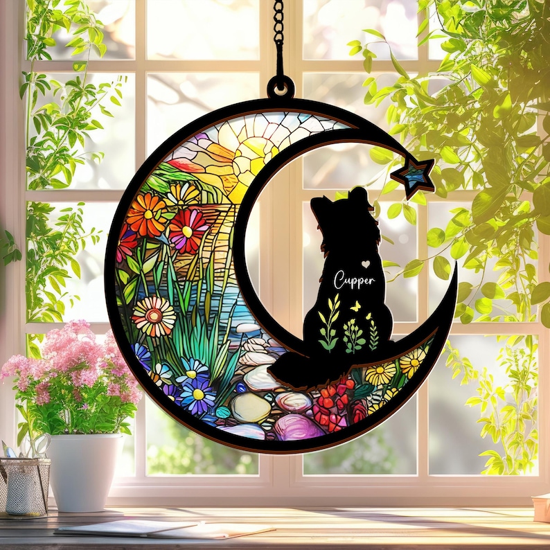 Puede incluir: Un carill&oacute;n de viento en forma de luna de vidrio tintado con una silueta de un perro sentado en la luna creciente. El vidrio presenta un dise&ntilde;o floral colorido con una salida del sol en el fondo. El nombre del perro, "Cupper", est&aacute; escrito en su pecho.