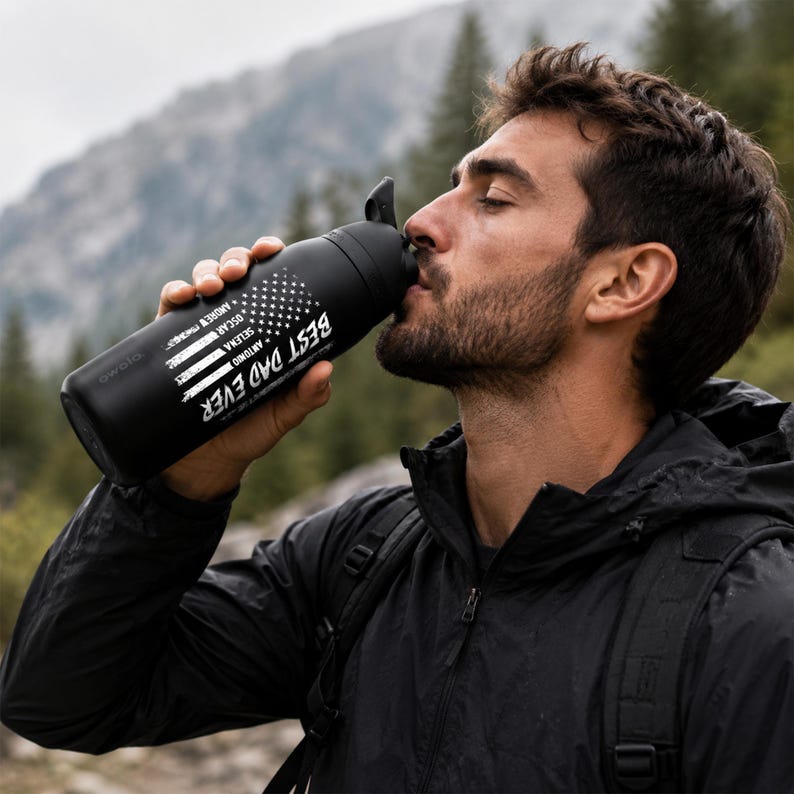 Puede incluir: Botella de agua negra con el texto "BEST DAD EVER" y un dise&ntilde;o de bandera estadounidense blanca y azul. La botella tambi&eacute;n tiene los nombres "ANDREW, OSCAR, SELENA, ANTONIO" impresos. La botella se sostiene cerca de la boca de una persona.