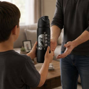 May include: A black water bottle with a silver eagle design and the word "DAD" printed on it. The bottle is being held by a child and offered to an adult. The bottle has a black lid and a silver arrow design.