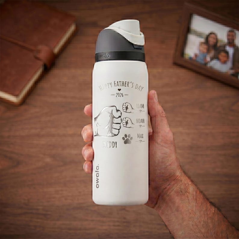 May include: A white Owala water bottle with a gray lid. The bottle has the text "Happy Father's Day - 2026" and a graphic of a fist, three smaller fists, and paw prints. The names Liam, Noah, and Max are below the smaller fists.