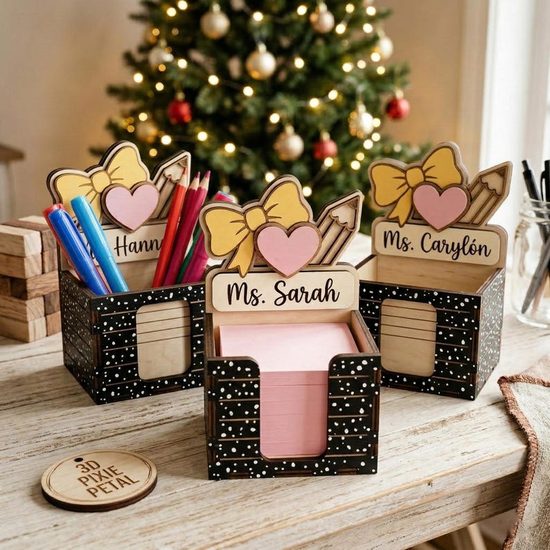 May include: Three wooden desk organizers with black and white speckled sides. Each has a bow, heart, and pencil design on top. One says "Hann", another "Ms. Sarah", and the third "Ms. Carylon". Pens, pencils, and pink sticky notes are inside.