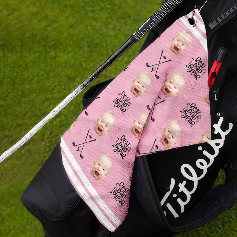 Op de afbeelding: Roze golfhanddoek met een herhalend patroon van een babygezicht en de tekst "Best Dad by far".