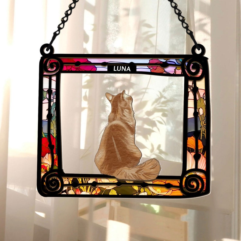 May include: A stained glass window hanging with a floral design and a cat looking out the window. The cat is brown and white and the name "LUNA" is written at the top of the window.