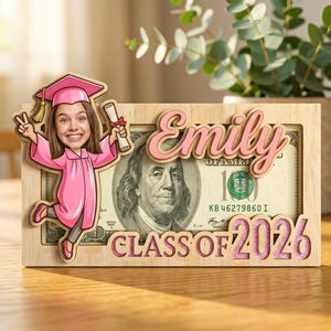 May include: A wooden graduation frame with a cartoon illustration of a graduate in a pink cap and gown. The frame includes the name "Emily" in pink and the text "CLASS OF 2026" with a dollar bill visible in the background.