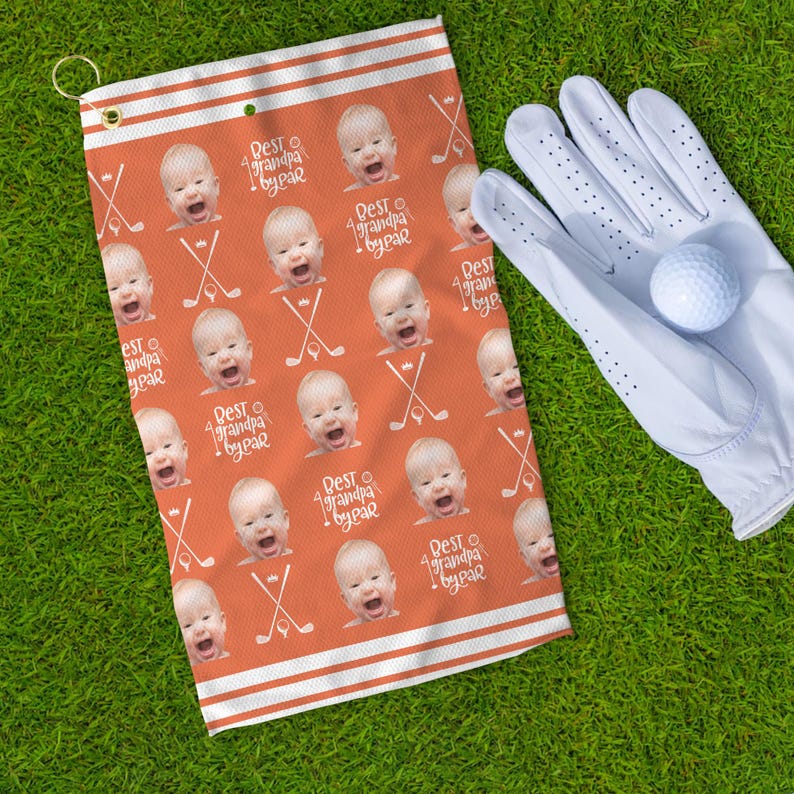 Op de afbeelding: Oranje en witte golfhanddoek met een herhalend patroon van een babygezicht en de tekst "Best grandpa by far".