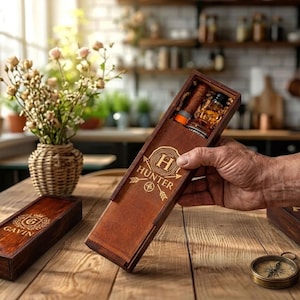 May include: A wooden gift box with the name "Hunter" engraved on the front, holding a cigar and a small bottle. The box is brown and has a sliding lid. A second box with the name "Gavin" is also visible. A compass is on the table.