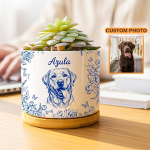 May include: A white ceramic planter with a blue line drawing of a Labrador Retriever and the name "Azula". The planter is filled with a green succulent and sits on a wooden base. A small photo of a dog is in the background.