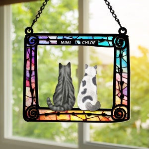 May include: A stained glass window hanging with two cats sitting side by side. The cats are facing away from the viewer. The window is decorated with a rainbow of colors and the names "Mimi" and "Chloe" are written at the top.
