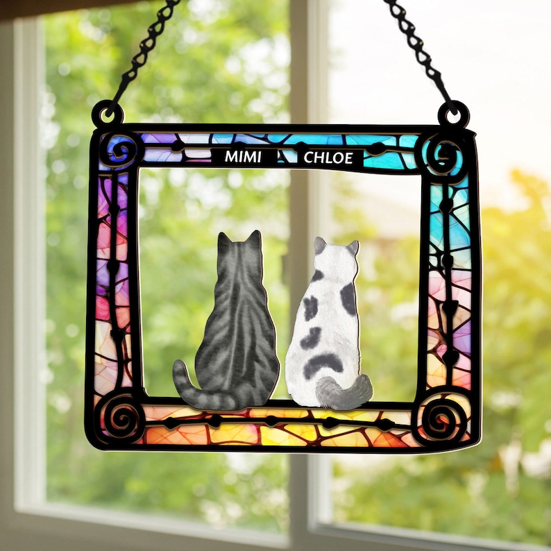 May include: A stained glass window hanging with two cats sitting side by side. The cats are facing away from the viewer. The window is decorated with a rainbow of colors and the names "Mimi" and "Chloe" are written at the top.