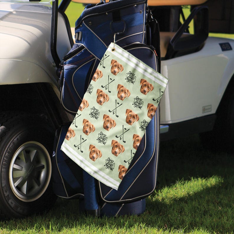 Op de afbeelding: Een groene en witte golfhanddoek met een herhalend patroon van een hondenkop en golfclubs. De tekst "Best Dad Ever" wordt herhaald op de handdoek. De handdoek hangt aan een golftas.