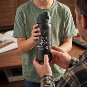 May include: A black water bottle with a flip-top lid. The bottle has white text that reads "Happy Father's Day 2026" and "Daddy". It also features a graphic of a fist and paw prints. The bottle is being held by two people.