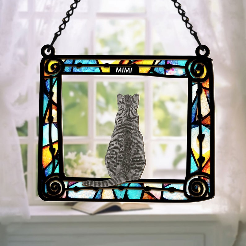May include: A stained glass window hanging with a black chain. The window is a rectangular frame with a colorful stained glass design. The window features a gray cat sitting inside the frame. The cat is looking out the window. The text "MIMI" is at the top of the frame.