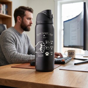 May include: Black Owala water bottle with a black lid. The bottle has white text that reads "Happy Father's Day - 2026" with a fist graphic and the names Liam, Noah, and Max. A paw print graphic is also included.