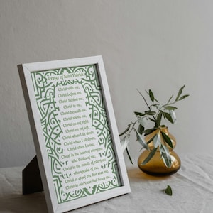 May include: A framed print of the "Prayer of Saint Patrick" in a white frame. The text is in green, with decorative Celtic knotwork around the edges. The frame is propped up on a dark brown stand. A small vase with greenery is in the background.