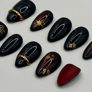 May include: Set of black and burgundy press-on nails with gold accents. The nails feature a glossy finish and various designs, including gold starbursts, curved lines, and ornate symbols. One nail is solid burgundy.