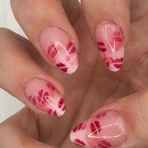 Könnte beinhalten: Nahaufnahme einer Hand mit langen, oval geformten Nägeln. Jeder Nagel ist mit einer hellrosa Basis und einem Muster aus roten Lippen bemalt. Die Nägel sind glänzend und gepflegt und zeigen ein Valentinstag-Thema.