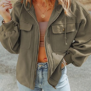 May include: An olive green hooded jacket with a full zipper closure, two front pockets, and drawstring details. The jacket is paired with blue jeans. The model is wearing gold necklaces and bracelets.