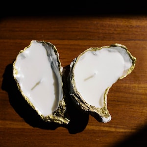 May include: Two oyster shell candles with white wax and a single wick. The shells have a gold-painted edge and sit on a dark wooden surface. The candles are a decorative home accent.