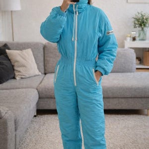 Puede incluir: Traje de esquí vintage azul claro con cuello alto y cremallera blanca. El mono de nieve de una pieza tiene una textura acolchada y presenta la marca "Stratos" en la parte superior del brazo. El traje se usa con calcetines blancos.