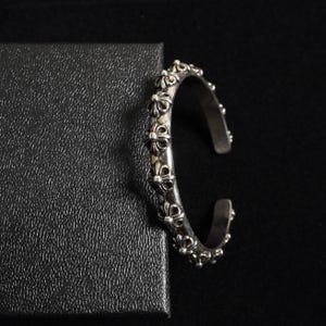 May include: A silver cuff bracelet with a floral design. The open-ended bracelet features a series of repeating, stylized flower motifs along its outer surface. The bracelet is set against a black background and a textured black surface.