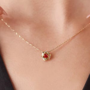 May include: A gold-colored necklace with a pendant featuring a red gemstone surrounded by small, clear stones. The pendant is shaped like a flower. The necklace is displayed against a skin-toned background.
