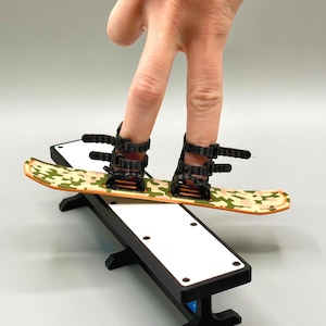 Puede incluir: Un mini snowboard con un patrón de camuflaje en verde, beige y naranja, con fijaciones negras. Se coloca sobre una rampa en blanco y negro, creando una escena deportiva y lúdica. La imagen muestra un artículo deportivo único a pequeña escala.