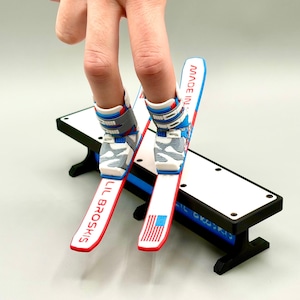 May include: Miniature skis with red, white, and blue detailing, with the text "LIL BROSKIS" and an American flag design. The skis are attached to a pair of fingers, resting on a black and white bench.